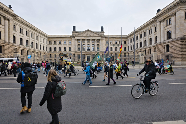 Eine Gruppe von Menschen geht vor einem großen Gebäude mit Fenstern, Säulen und Bögen die Straße entlang, einige tragen Taschen und Fahnen, andere fahren Fahrräder, im Hintergrund sind Fahrzeuge und Laternenmasten zu sehen.