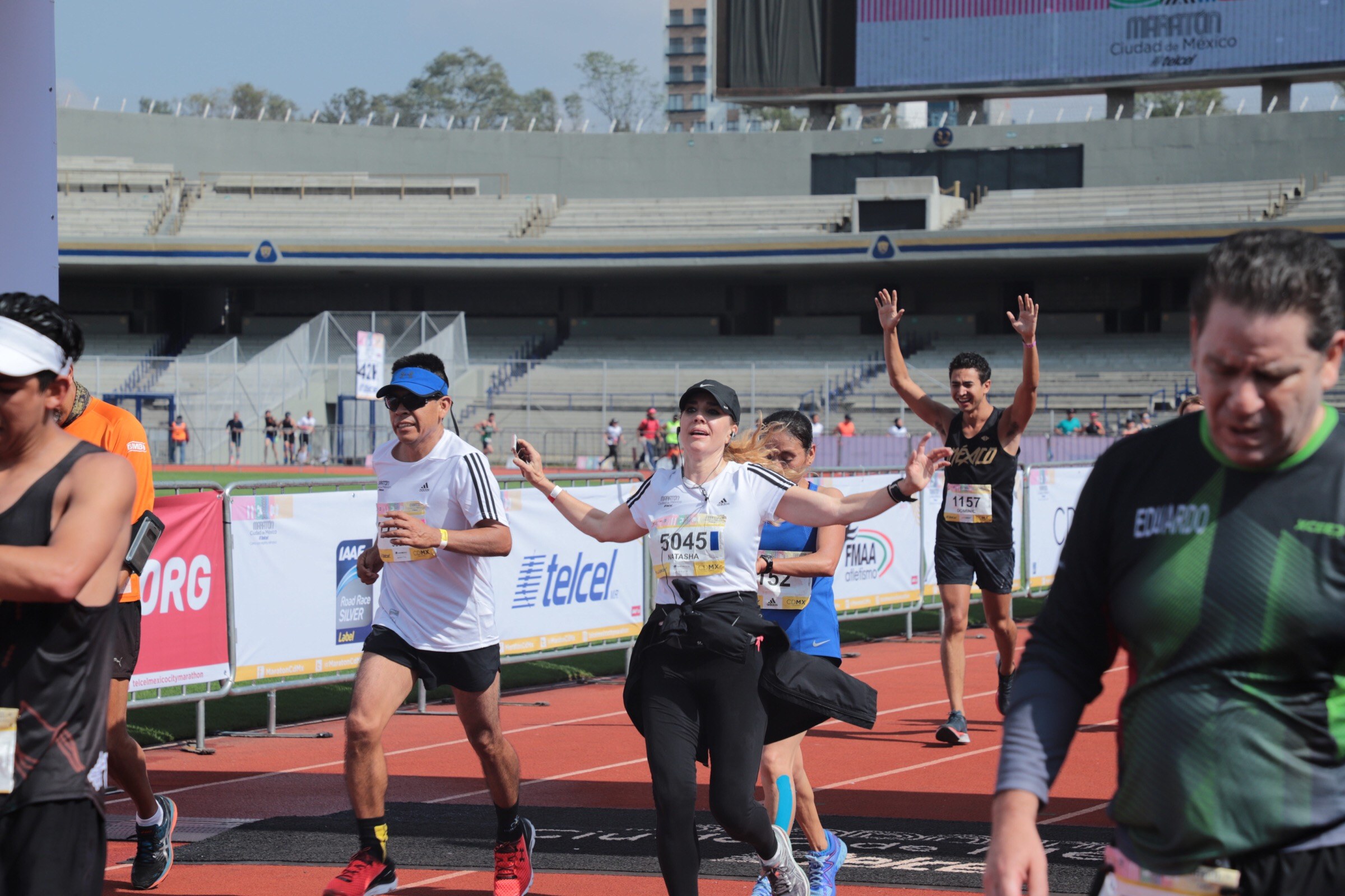Gruppe von Läufern auf einer Stadionbahn mit Bannern, einem Display, einem Gebäude, Bäumen und einem bewölktem Himmel; einige Läufer tragen Turnschuhe und Hüte.