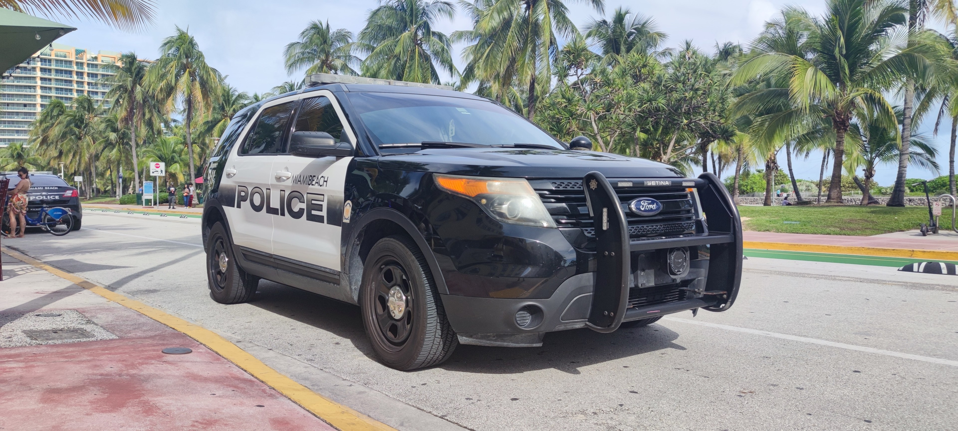Polizeiauto am Straßenrand mit Menschen, Fahrrädern, Gras, Bäumen, Gebäuden und einem klaren blauen Himmel im Hintergrund; Vorderrad sichtbar.