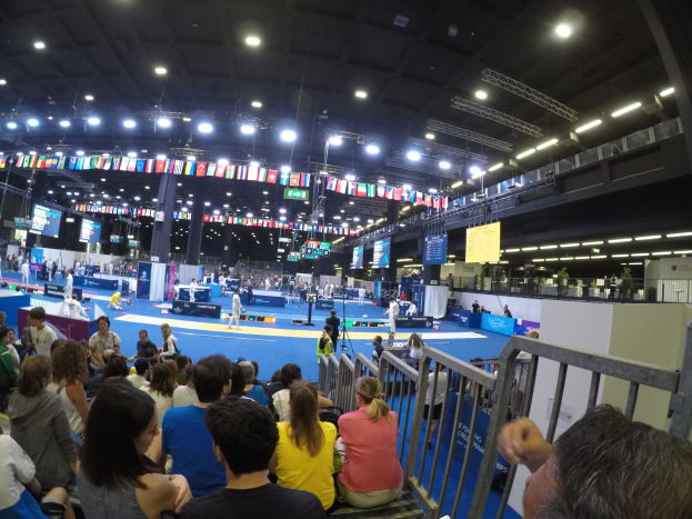 Eine große Menschenmenge sitzt einer Bühne mit einem Geländer auf der rechten Seite und einer Decke mit Lampen gegenüber, während ein paar Menschen auf der Bühne stehen.