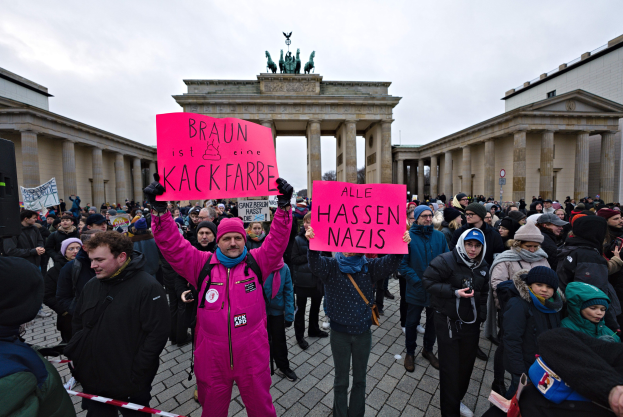 Eine Gruppe von Menschen demonstriert vor dem Brandenburger Tor in Berlin, hält Schilder gegen die Anti-Nazis-Bewegung der deutschen Regierung und zeigt Gebäude, Säulen und Statuen im Hintergrund unter einem klaren blauen Himmel.
