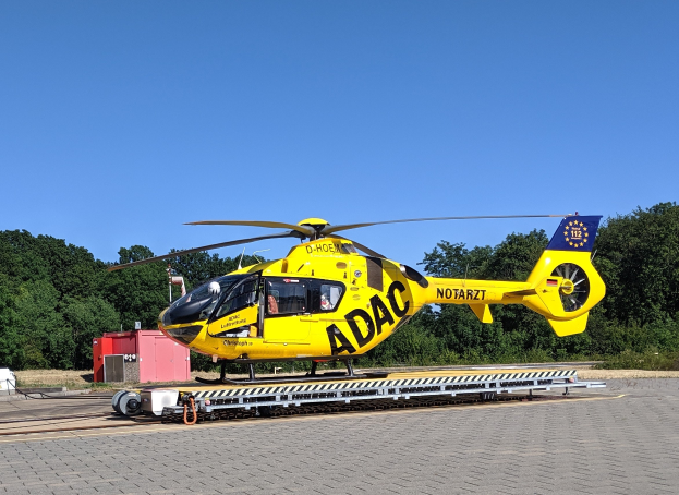 Ein gelber Hubschrauber sitzt auf einem Flughafen-Asphalt, umgeben von Bäumen und einem klaren blauen Himmel, mit verschiedenen Gegenständen in der Nähe des Hubschraubers.