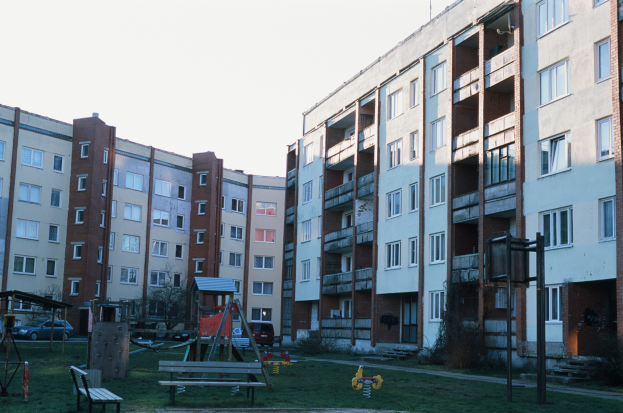Wohngebäude mit Spielplatz, Rasenfläche, Bänken, Schaukeln, Fahrzeugen, Bäumen und einem klaren blauen Himmel im Hintergrund.