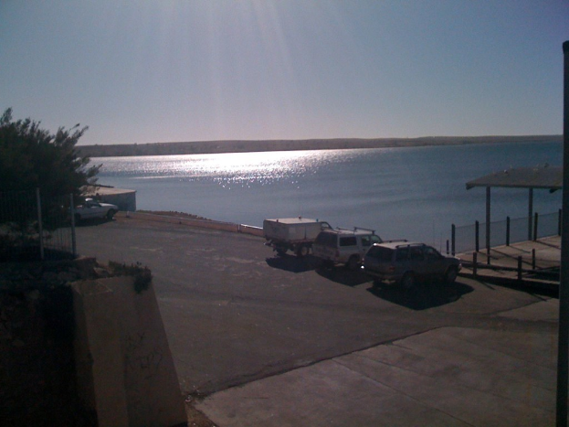 Autos fahren auf einer Straße mit Bäumen links, einem Unterstand rechts und Wasser im Hintergrund.