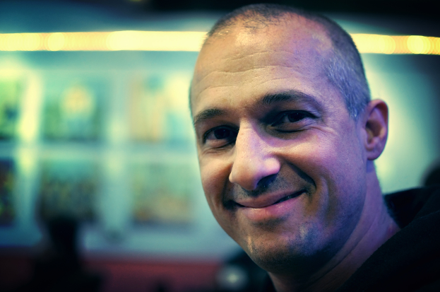 Ein Mann in einem schwarzen Kleid l├Ąchelt mit einem unscharfen, hell erleuchteten Hintergrund.