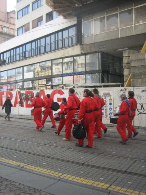 Menschen, die vor einer Zaunwand mit Plakaten und Texten spielen und gehen, hinter der es Gebäude mit Glaswänden und Fenstern gibt, auf denen Namen stehen.