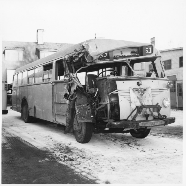 Schwarz-weiß-Foto eines schwer beschädigten Busses am Straßenrand mit eingedellter Front und zersplitterten Fenstern neben Gebäuden.