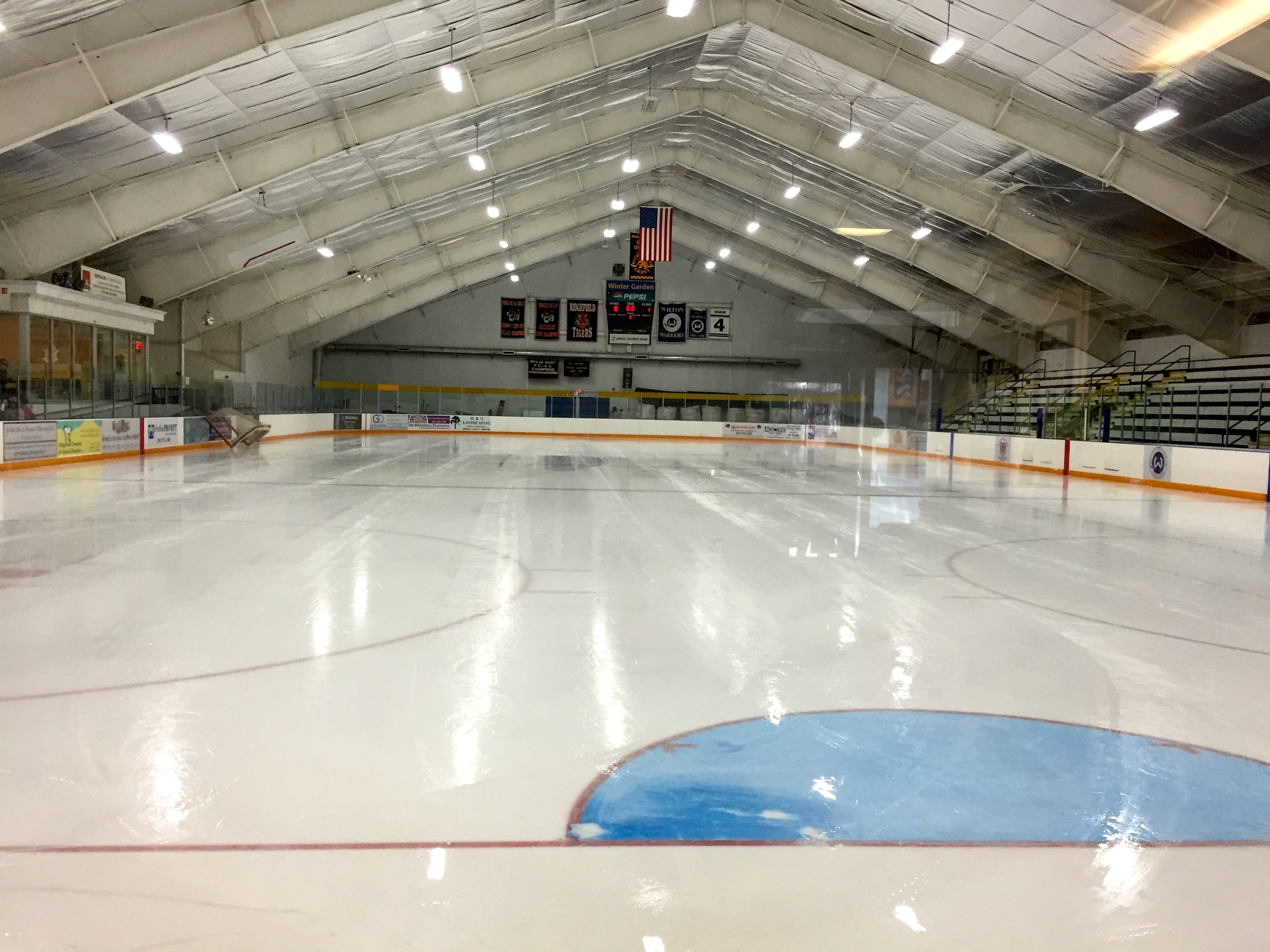 Große Indoor-Eisbahn mit Deckenbeleuchtung, Werbetafeln, einer Wand, Treppen mit Geländern und einigen Menschen, die eine Sportanlage anzeigen, die likely für Hockey verwendet wird.