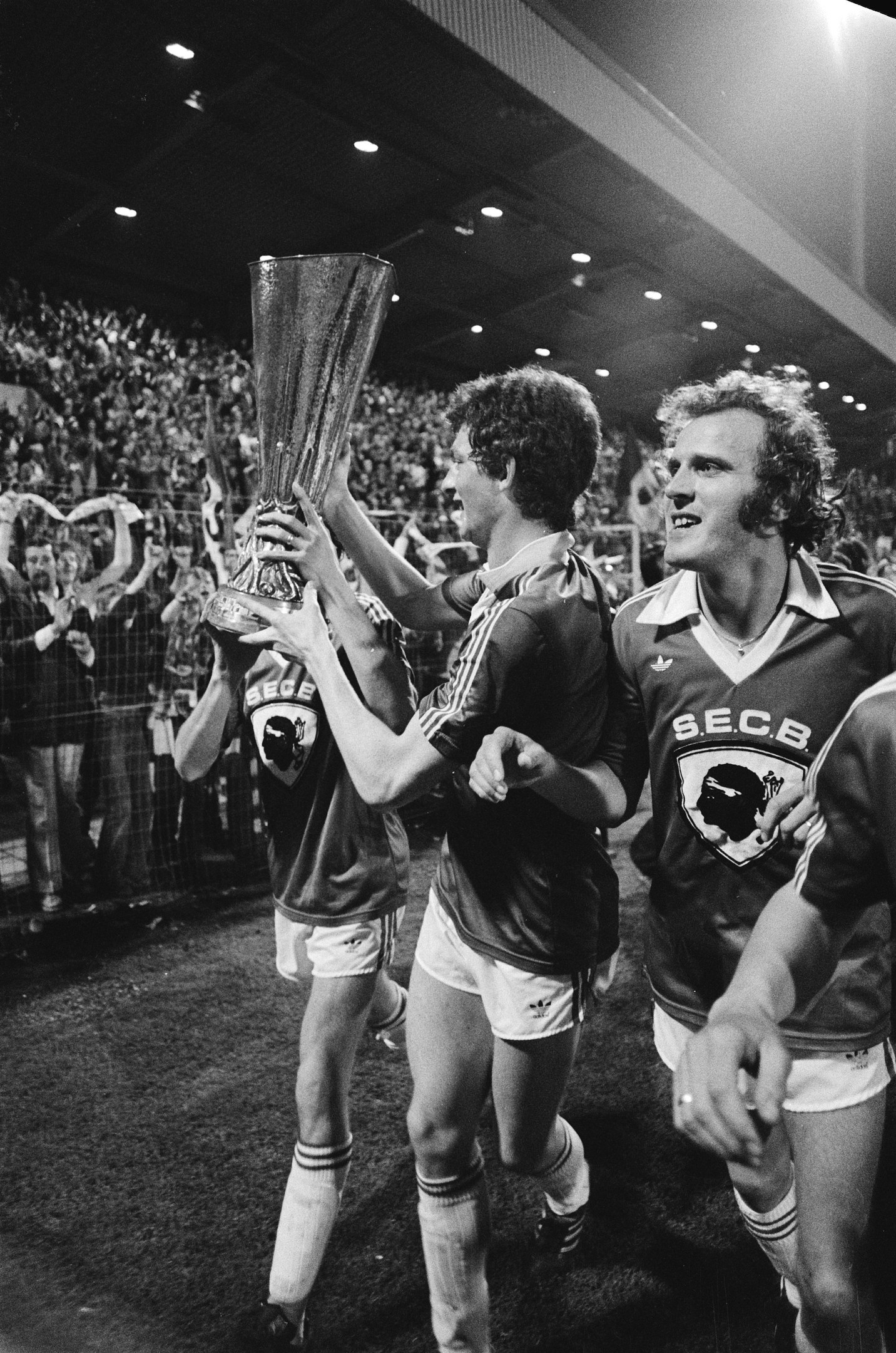 Schwarzes und weißes Foto von Fußballspielern, die einen Pokal auf einem Feld halten und feiern, während Zuschauer hinter einem Zaun zusehen.