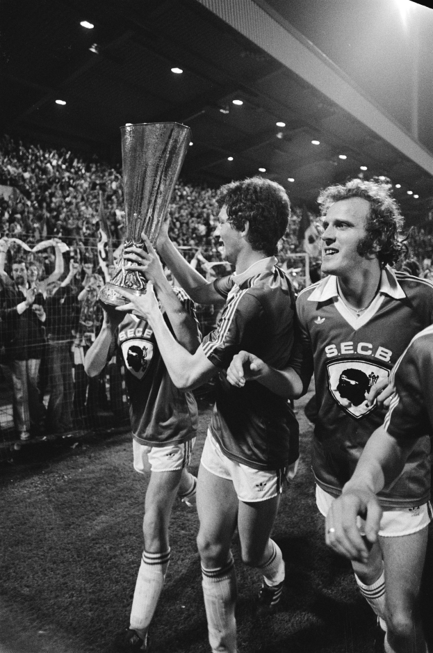 Schwarzes und weißes Foto von Fußballspielern, die einen Pokal auf einem Feld halten und feiern, während Zuschauer hinter einem Zaun zusehen.