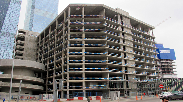 Großes Bauprojekt in einer Stadt mit Hochhäusern, Fahrzeugen, Fußgängern, Verkehrsinfrastruktur und Baumaschinen vor einem klaren Himmel.