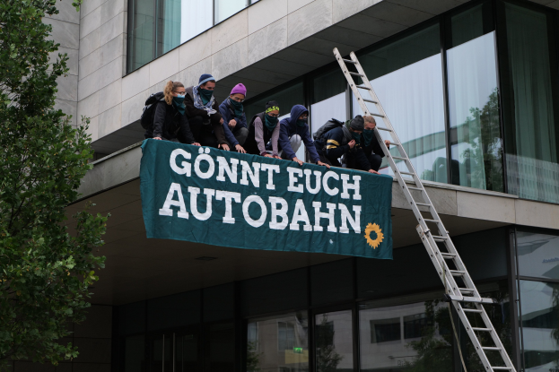 Eine Gruppe von Menschen auf einem Gebäude mit einer Fahne, einem Baum auf der linken Seite und Glasfenstern im Hintergrund, die scheinbar für die deutsche Regierung protestieren.