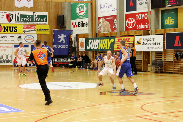 Ein Basketballfeld mit Spielern im Vordergrund und einer hölzernen Wand mit Postern und Bannern im Hintergrund.