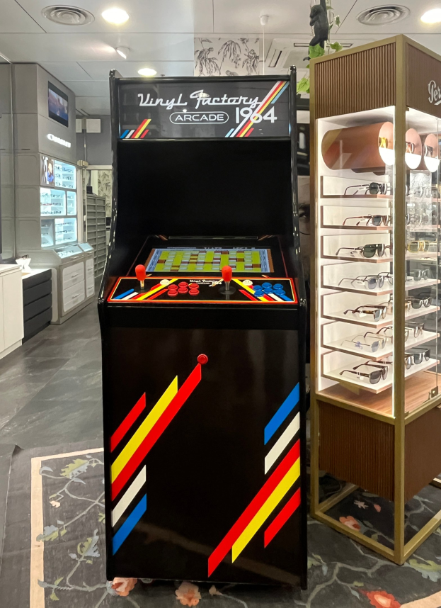 Ein Ladengeschäft mit einem zentralen Vintage-Arcade-Automaten, einem Gläserregal auf der rechten Seite und Hintergrund-Elementen wie Schäubern, einem Fernseher und anderen Gegenständen, mit Deckenlampen, die für eine Retro-Atmosphäre sorgen.