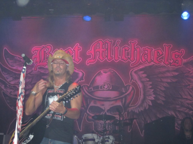 Eine Konzertszene mit einem Gitarristen links neben einem Mikrofon, einer Person in der unteren rechten Ecke, einem Banner im Hintergrund und Drogen in der Mitte unten, mit Licht an der Spitze.