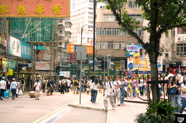 Eine belebte Outdoor-Szene mit Menschen auf einer Straße, umgeben von Pfählen, Signalen, Plakaten, Pflanzen und einem Baum, mit Gebäuden, Rohren und einem Plakat im Hintergrund.