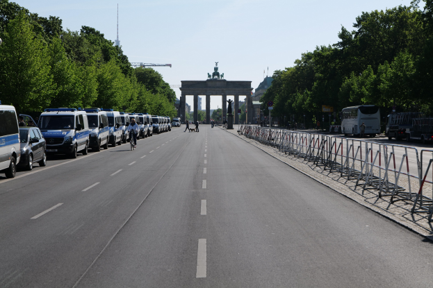 Lange Reihe von Polizeiwagen, die an einer Straße vor dem Brandenburger Tor geparkt sind, mit Menschen auf Fahrrädern und Fütgängern, Barrieren, Bäumen und einem Bogen mit Statuen im Hintergrund.