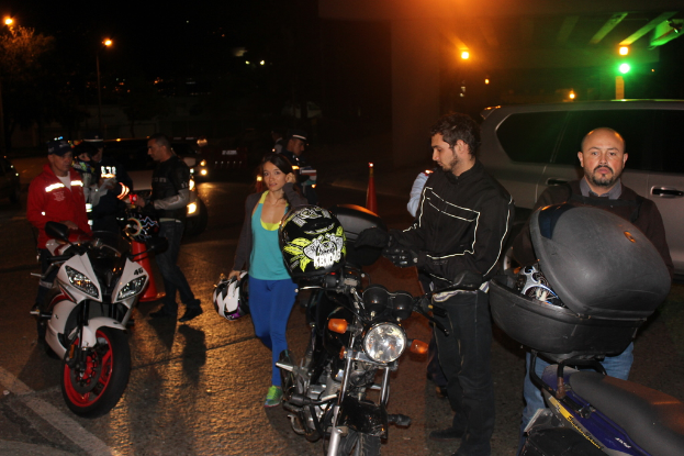 Eine Gruppe von Menschen steht und geht auf einer dunklen Straße in der Nacht, hält Gegenstände, mit einem teilweise sichtbaren Auto auf der rechten Seite, Straßenlaternen, Bäumen und einer Wand mit Lichtern im Hintergrund.
