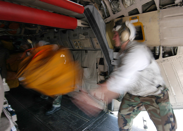 Ein Mann in weißem Hemd und Tarnkleidung, der einen Helm trägt, lädt eine gelbe Tasche in ein Flugzeug, umgeben von anderen mit Helmen, mit Gegenständen im Flugzeug, die auf einen Verletzten hindeuten.