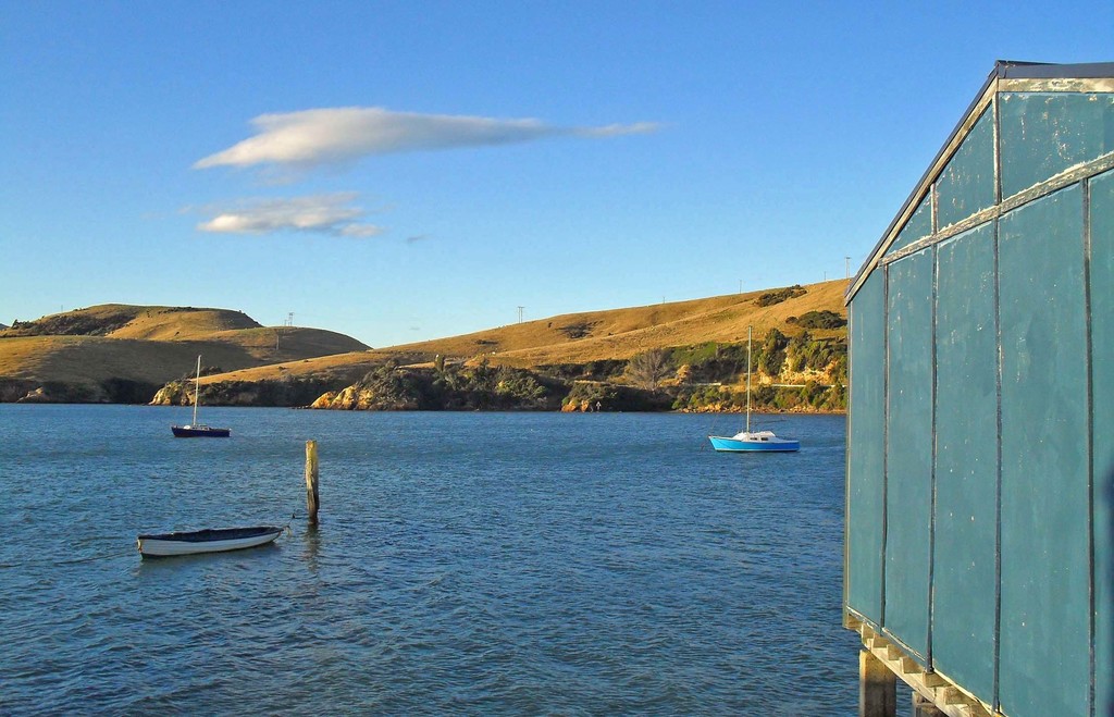 Boote und Pfähle im Wasser, ein Holzhaus rechts, Bäume und Berge mit Windmühlen im Hintergrund und Wolken am Himmel.