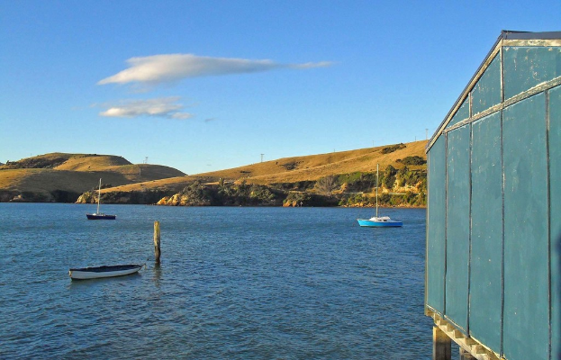 Boote und Pfähle im Wasser, ein Holzhaus rechts, Bäume und Berge mit Windmühlen im Hintergrund und Wolken am Himmel.