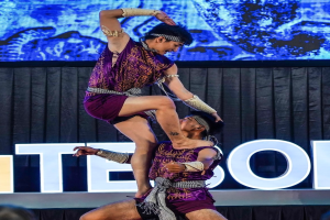 Ein Mann und eine Frau in lila Kleidern führen Akrobatik auf der Bühne vor, wobei der Mann die Frau in seinen Armen hält, während das Publikum unten zusieht und ein schwarzer Vorhang und ein Bildschirm im Hintergrund zu sehen sind.