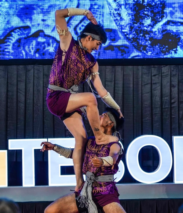 Ein Mann und eine Frau in lila Kleidern führen Akrobatik auf der Bühne vor, wobei der Mann die Frau in seinen Armen hält, während das Publikum unten zusieht und ein schwarzer Vorhang und ein Bildschirm im Hintergrund zu sehen sind.