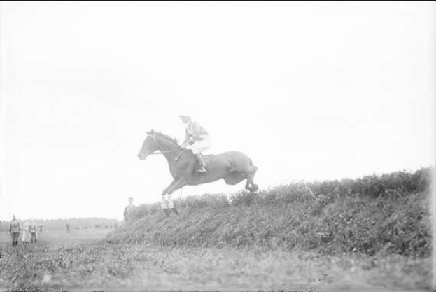 Ein Schwarz-Weiß-Foto eines Pferdes und seines Reiters, die über einen grasbewachsenen Hügel springen, wobei das Pferd seine Mähne und seinen Schweif im Wind wehen lässt und der Reiter einen Helm und einen langen Mantel trägt, während einige Menschen im Hintergrund zuschauen.