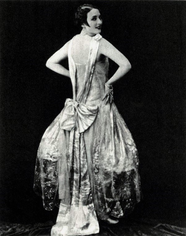 Eine Frau in einem Kleid im Stil der 1920er Jahre mit hohem Kragen, langen Ärmeln und einem weiten Rock posiert selbstbewusst mit einer eleganten Hochsteckfrisur und dezentem Make-up.
