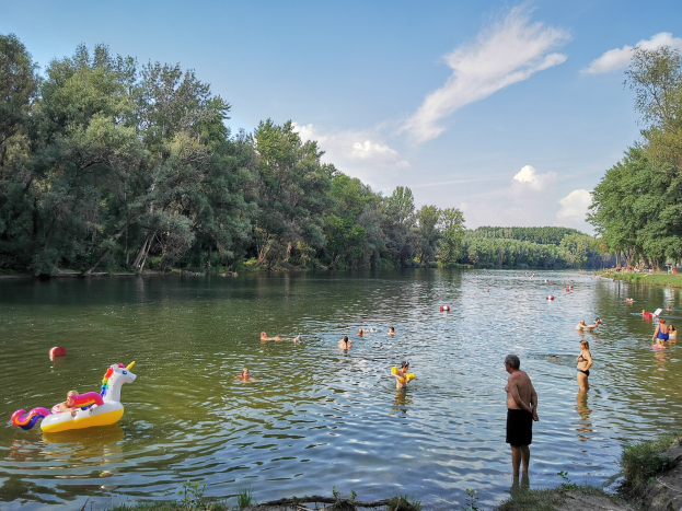 Menschen, die in einem See schwimmen, umgeben von grünem Gras und Bäumen unter einem blauen Himmel mit Wolken, eine Person steht am Ufer, und aufblasbare Spielzeuge treiben im Wasser.