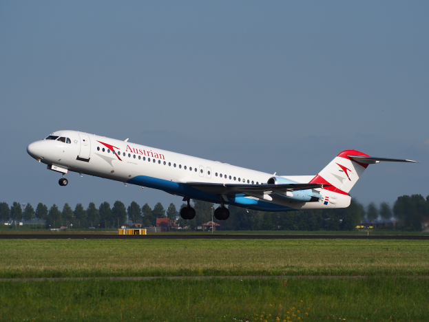 Ein Austrian-Airlines-Embraer-ERJ-190-Flugzeug beim Abheben vom Frankfurter Airport, mit gr√ľnem Gras, B√§umen und Geb√§uden im Hintergrund und dem Himmel √∂ber dem Flugzeug.