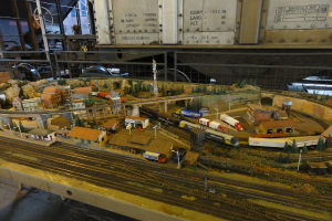 Eine Miniaturszene auf einem Tisch mit einer Schienenstrecke, einem Zug, Gebäuden, Fahrzeugen, einer Brücke, einem Turm und Bäumen.