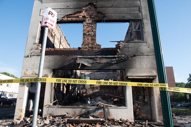 Ein abgerissener Bau inmitten einer Straße, umgeben von gelbem Absperrband, mit Schutt drumherum und anderen Gebäuden, Fahrzeugen, Bäumen und einem klaren blauen Himmel im Hintergrund.