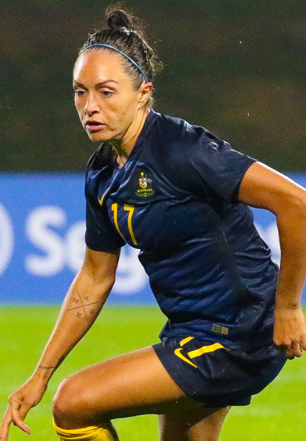 Eine Fußballspielerin im Einsatz auf dem Feld, die ein blaues T-Shirt und gelbe Shorts trägt, vor einem blauen Schild mit Text im Hintergrund.