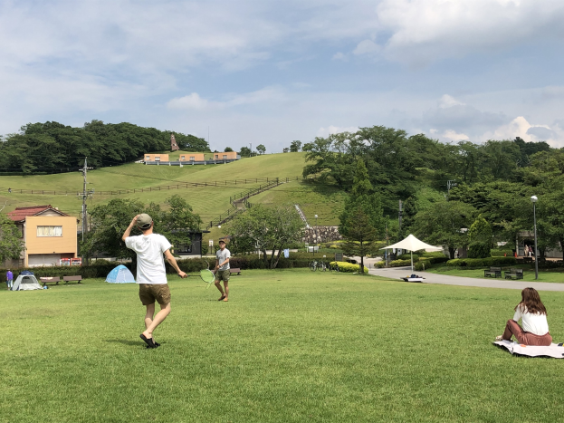 Menschen spielen Badminton in einem Park mit Zelten, Straßeneinrichtungsgegenständen und Gebäuden im Hintergrund.