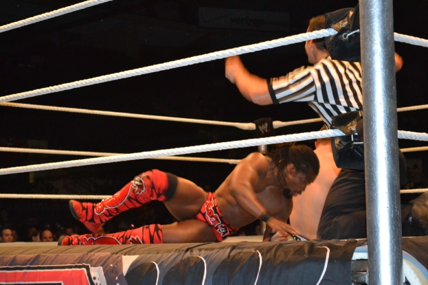 Zwei Männer kämpfen in einem Ring, einer trägt ein rotes Hemd und Schuhe, der andere kurze Hosen, mit einem Schiedsrichter rechts daneben und einer Zuschauermenge im Hintergrund unter einem dunklen Himmel.