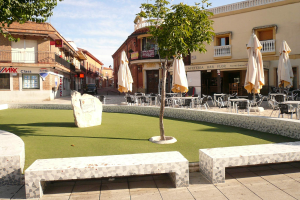 Ein Minigolf-Platz in einem Stadtplatz, umgeben von Bänken, Tischen, Stühlen, Sonnenschirmen, Bäumen und Gebäuden mit Fenstern, Geländern und Namensschildern.