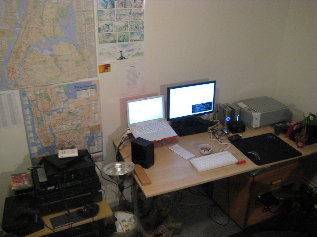 Ein Raum mit einem Tisch, auf dem ein Laptop, ein Lautsprecher, ein Monitor, eine Tastatur, ein Ball, eine Maus, ein Scanner und Papiere liegen; ein weiterer Tisch mit Elektronikgeräten ist links unten zu sehen, mit Charts und Postern an der Wand dahinter.