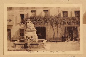 Ein altes Foto eines Brunnens mit einer Statue auf einem Sockel in der Mitte, umgeben von Menschen auf Bänken, mit einem Baum im Hintergrund und Text unten.