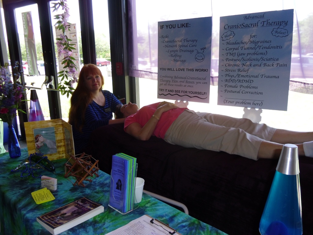 Eine Frau liegt auf einer Therapieliege, während eine andere Frau eine Therapiesitzung durchführt, mit Postern, die die Therapie beschreiben, und einem Tisch mit Gegenständen und Büchern daneben.