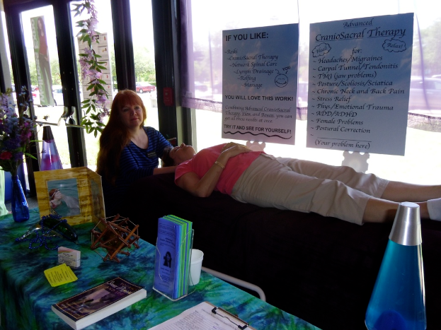 Eine Frau liegt auf einer Therapieliege, während eine andere Frau eine Therapiesitzung durchführt, mit Postern, die die Therapie beschreiben, und einem Tisch mit Gegenständen und Büchern daneben.