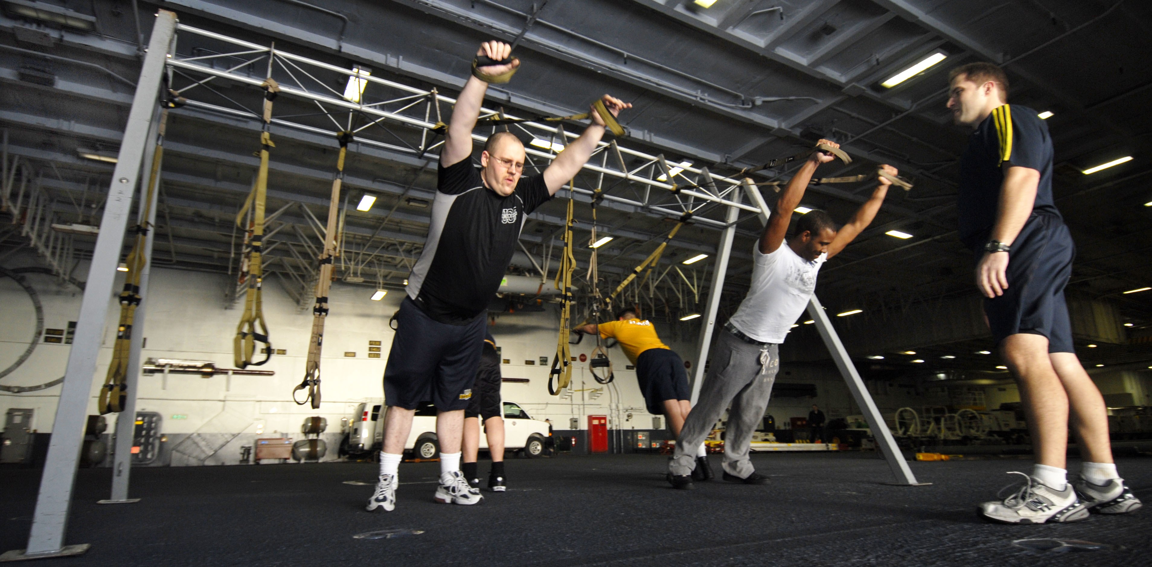 Mannergruppe beim Pull-up an Metallstangen in einem Fitnessstudio mit Deckenbeleuchtung und diversen Gegenständen im Hintergrund, ein Mann in der Mitte mit Schuhen.