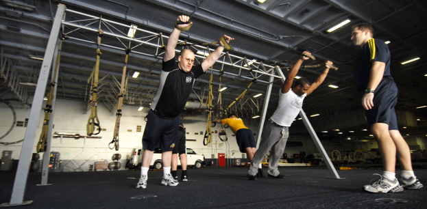 Mannergruppe beim Pull-up an Metallstangen in einem Fitnessstudio mit Deckenbeleuchtung und diversen Gegenständen im Hintergrund, ein Mann in der Mitte mit Schuhen.