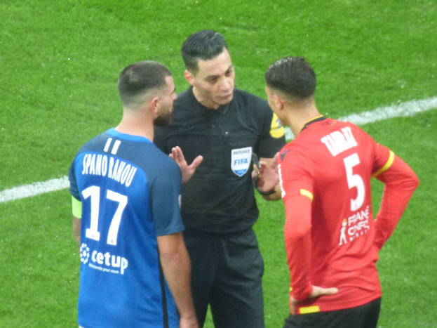 Schiedsrichter spricht mit zwei Männern auf einem Fußballfeld, einer in schwarzer Hose und Hemd, der andere in einem blauen und roten T-Shirt mit Text und Brille.