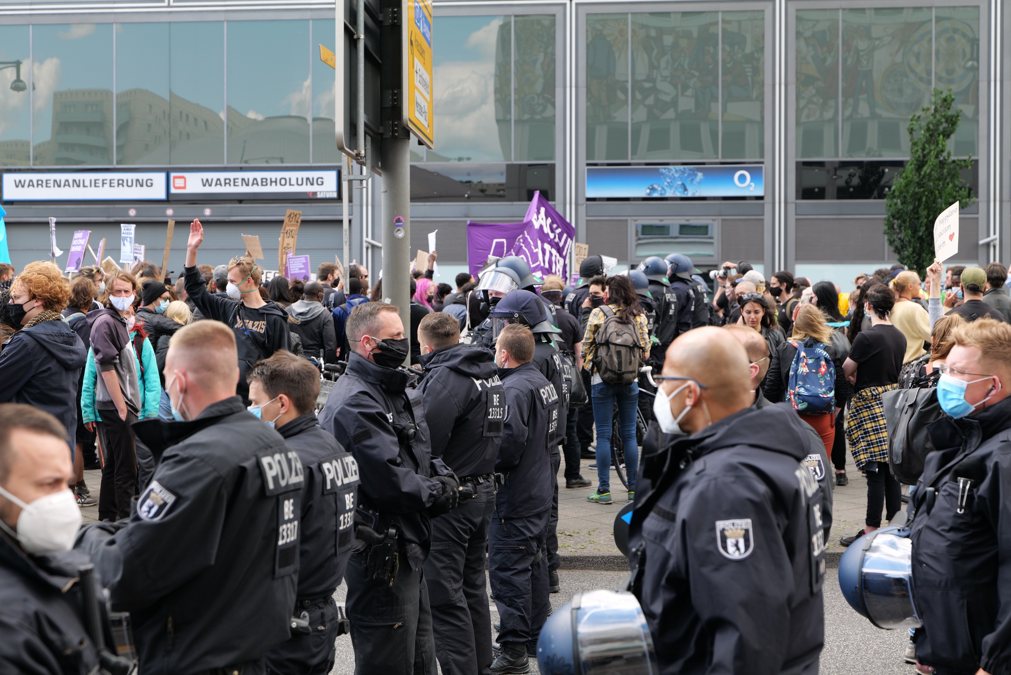 Große Menschenmenge vor einem Gebäude protestierend, einige halten Schilder und tragen Helme, mit einem Pfahl und einem Schild im Vordergrund und einem Baum im Hintergrund.