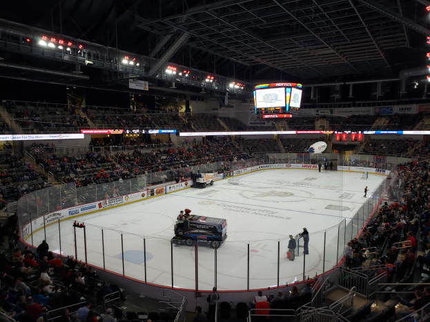 Ein Eishockeyspiel in einer großen Arena mit Fahrzeugen auf dem Eis, Zuschauern um die Bahn und Menschen auf den Tribünen, sowie Bretter, Lampen und ein Display im Hintergrund.