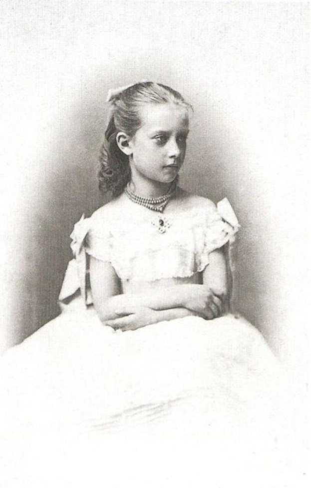 Schwarzes und weißes Foto eines jungen Mädchens in einem weißen Kleid, das angeblich die Zarin Alexandra Fjodorowna von Russland ist, die eine zarte Kette trägt und ihr Haar ordentlich hochgesteckt hat und einen majestätischen, würdevollen Gesichtsausdruck hat.