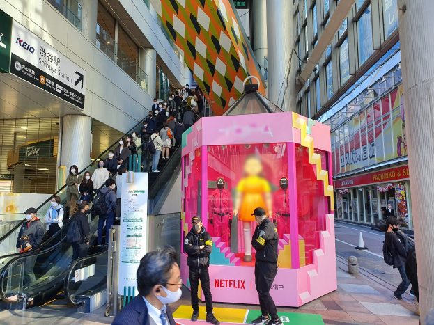 Eine Gruppe von Menschen steht vor einem Gebäude mit Glaswänden, Säulen und Texttafeln, einige tragen Masken, während hinter ihnen eine Rolltreppe mit Menschen nach oben führt; ein pinker Kasten in der Mitte zeigt Schaufensterpuppen und trägt die Aufschrift "Netflix's new Netflix booth in Tokyo."