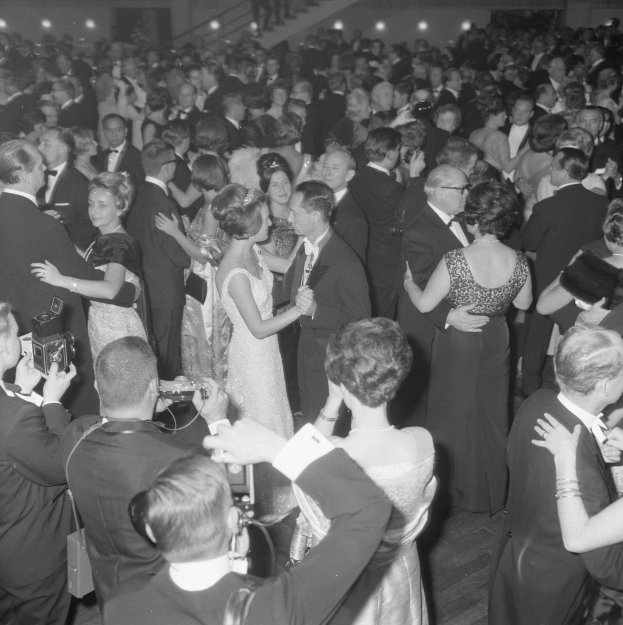 Ein Schwarz-Weiß-Foto einer Gruppe von Menschen, die auf einer Tanzfläche tanzen, einige halten Kameras in den Händen, mit Treppen und Lichtern im Hintergrund.