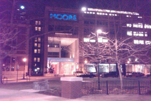 Ein Krankenhausgebäude mit Fahrzeugen, Bäumen und einem Zaun davor.